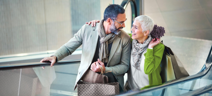 Man and woman travelling up escalator in a mall with shopping bags in hand smiling at eachother